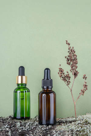 Two brown, green glass bottles with serum, essential oil or other cosmetic product and dry flower plant on tree bark covered with moss against green background. Natural Organic Spa Cosmetic concept.の写真素材