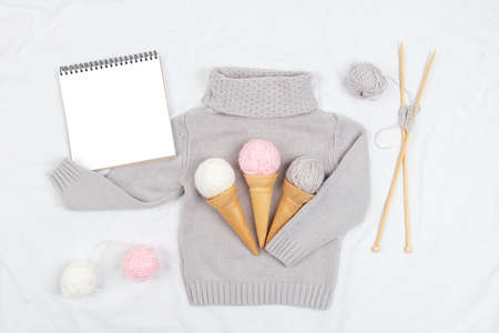 Three ice cream from yarn and waffle cones on a gray knitted sweater, wooden knitting needles and open notepad on white background. Knitting, hobby and DIY concept. Top view Flat lay.の写真素材
