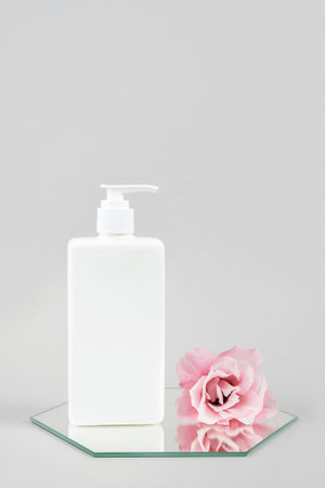 White cosmetic blank bottle with dispenser and pink flower on mirror, gray background. Natural Organic Spa Cosmetic Beauty Concept Mockup, Front view.の写真素材