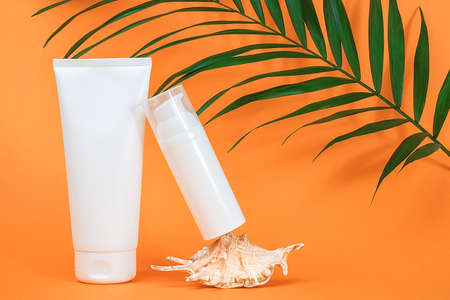 Two white blank cosmetics bottles with sunscreen, suncream or other cosmetic product, seashell and green brunch palm on orange background. Concept skin care in summer holiday Mockup Front view.の写真素材