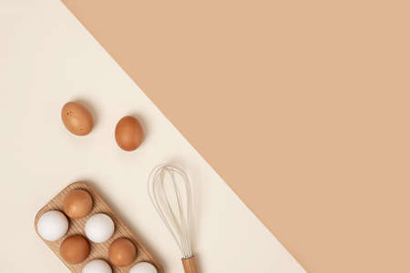 Eggs in wooden egg box and whisk on beige background. Flat lay Top view Copy space.
