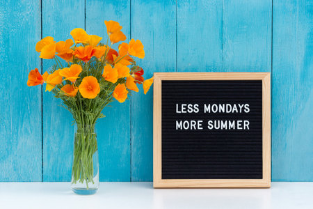 Less mondays more summer. Motivational quote on letter board and bouquet orange flowers on white table against blue wooden wall. Concept inspirational quote of the day. Front view.の写真素材