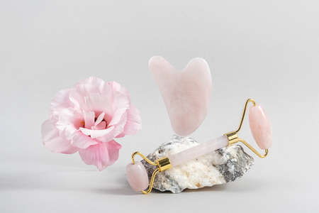 Crystal rose quartz facial roller and massage tool jade Gua sha on stones and natural pink flower on...