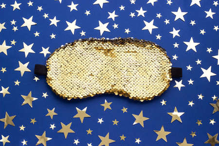 Shiny sleep mask for eyes decorated with sequins and gold stars confetti on a blue background. Top...