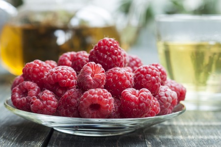 Fresh picked raspberries on a background of green tea with hedgehogs in a glass transparent teapot on an old wooden table. A natural herbal drink.の写真素材