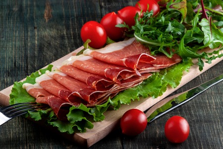 On the table is a wooden cutting board with sliced jamon, tomatoes and herbsの写真素材