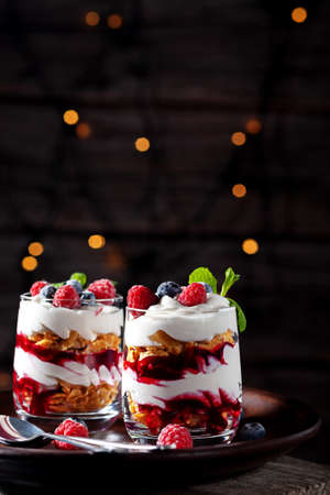 On a wooden background with lights, dessert with cream, cottage cheese, raspberries and blueberries in a glassの写真素材