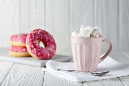 Donuts with pink glaze and colored caramel powder and cocoa with marshmallows on a white wooden...
