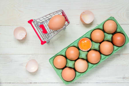 top view of raw brown chicken eggs in green egg carton box and shopping cart on white wooden...