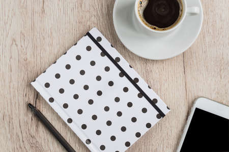 Business and office concept - black and white polka dot cover notebook, smartphone and cup of black coffee on wooden table. top view.の写真素材