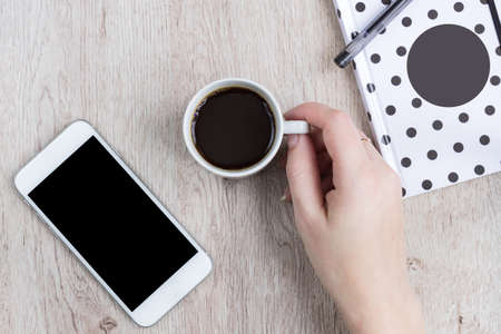 Business and office concept - black and white polka dot cover notebook, smartphone and cup of black coffee on wooden table. top view.の写真素材