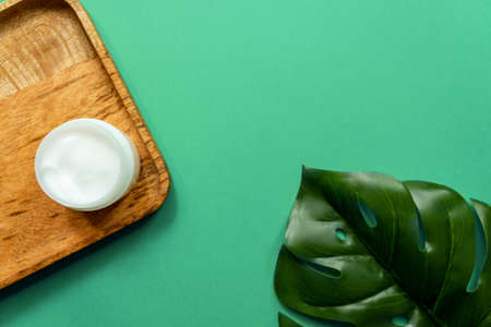 White jar of face cream on a wooden tray. green background, top view, tropical leaves. Concept of...