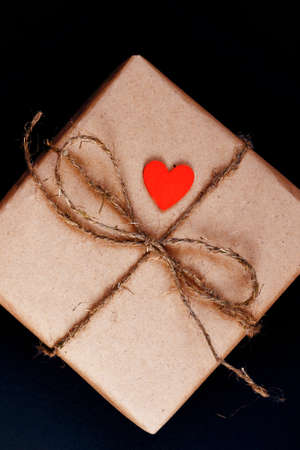 Handcrafted gift box wrapped in Craft paper with red wooden heart, rope and bow on black background. Top view, flat layの写真素材