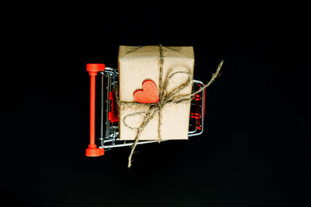 Handcrafted gift box wrapped in Craft paper with red wooden heart and rope in toy shopping cart, on black background. Top view, flat layの写真素材