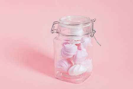 White and pink twisted meringues in a glass jar on pink background. French dessert prepared from...