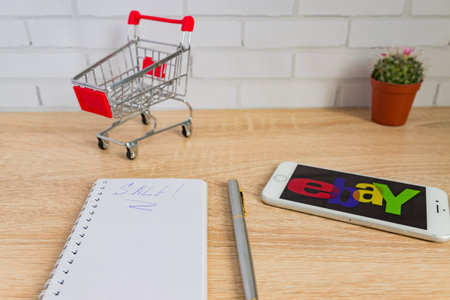 Moscow, Russia - March 13, 2019: Close up of ebay app on a Apple iPhone 8 screen, shopping cart and notebook with sale handwritten word. shopping online. international electronic commerce company.のeditorial素材