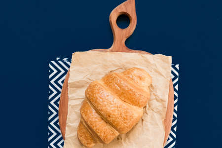 top view of croissant on cutting board and craft paper on blue background with copy space.