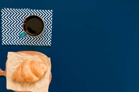 top view of cup of black coffe and croissant on a cutting board and craft paper on blue background with copy space. Morning breakfast in french style.の写真素材