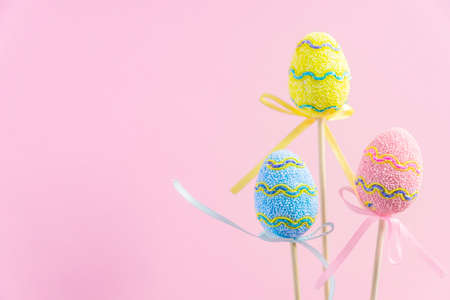 Easter purple, yellow and blue decorated eggs stand on a wooden sticks on pink background. Minimal...