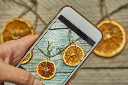 woman takes a photo of christmas decoration made of dried orange slices on her white modern smarthfone. Taking blogging conent for christmas and new yearの写真素材