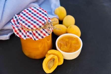 Glass jar with homemade organic apricot jam and ripe apricots on black background. Rustic style,...