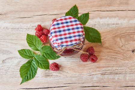 fresh raspberry jam in a glass jar on a wooden table, next to fresh raspberries with green leaves....
