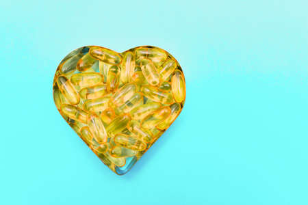 Fish oil capsules in a heart shape plate on blue background, vitamin D supplement, top view with...