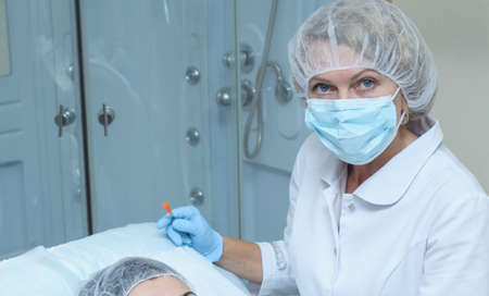 Portrait of middle aged woman doctor cosmetician in white lab coat, mask and sterile gloves makes...