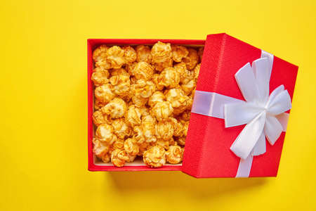 Red gift box with white bow full of tasty caramel popcorn on yellow background. Top viewの写真素材