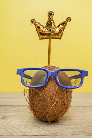 Funny coconut in glasses with sequin crown on yellow background. Summer and travel conceptの写真素材