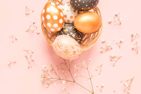 Top view of wooden painted eggs in gold, black and rose colors in wicker basket with branch of gypsophila on pink background. Happy easter backgroundの写真素材