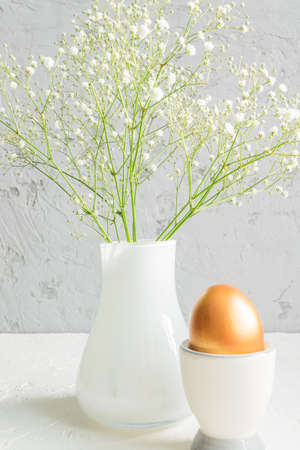 Close up of Golden egg in egg cup on the background of bouquet of gypsophila. Cozy easter still life