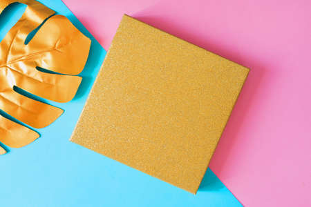 Top view of golden gift box and monstera leaf on double pink and blue background. Minimal flat lay,...