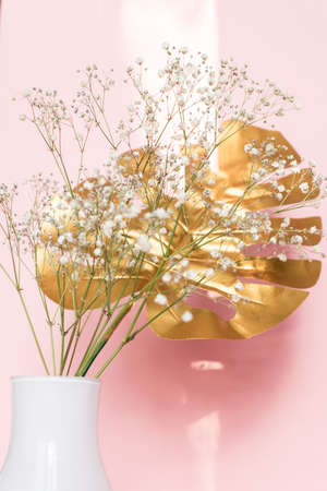 White Vase with golden tropical leaf and gypsophila flowers against pink background. Stylish interior decoration.の写真素材