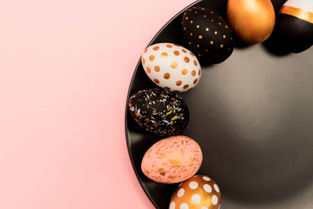 Top view of pink, white and golden decorated eeaster eggs on black plate on pink background. Trendy...