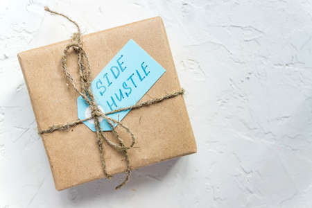 Side hustle written on blue tag against gift box wrapped in recycled paper background.