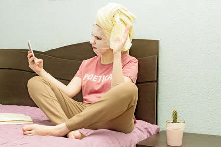 Caucasian cute young girl female woman sitting in bed with spa face care sheet mask doing selfie at...