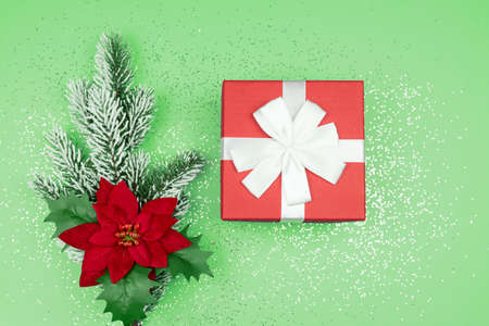 Top view of red gift box and fur tree branch with poinsettia on green background with confetti....