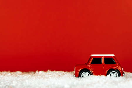 Small red toy car drives on a snowy road on red background. Festive christmas greeting card with...