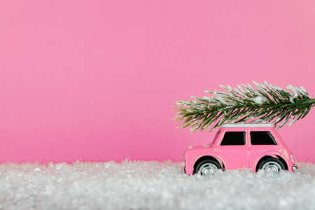Small pink toy car carrying spruse tree on a snowy road. Festive christmas greeting cardの写真素材