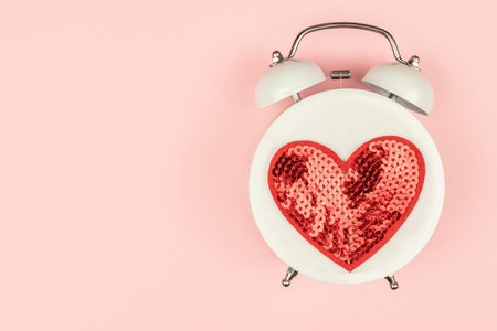 Pink Background with white alarm clock with red sequin heart. Creative layout. Top view. Time for love and greetingsの写真素材
