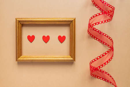 Valentine's day or Wedding romantic concept. Twisted decorative ribbon and golden photo frame with red hearts on beige backgroundの写真素材