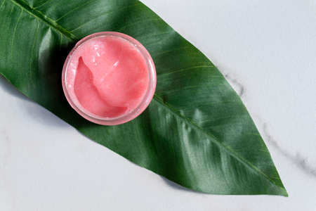 Pink body scrub on tropical leaf on marble background. Wellness and spa concept. Body care and...