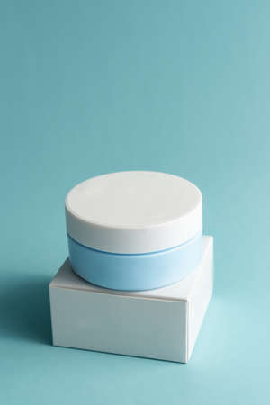Face cream jar with plastic cap and whire carton box, package on blue background. Mockup unbranded cosmetology product.の写真素材