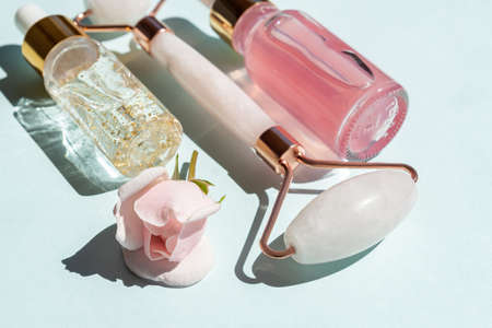 Massage roller for the face made of rose quartz with bottles of cosmetic oil or serum on a blue background. The concept of skin care at home. 24k gold serum and rose water for face treatmentの写真素材