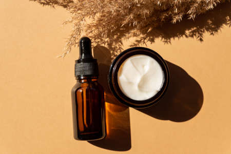 Moisturizer cream jar, serum in amber glass bottle and dry flowers reeds on beige background. Flat lay, top view. Set for skin and body care beauty products.の写真素材