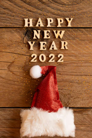 Happy new year 2022 wooden letters quote with red sparkling santa hat on wooden table with glitter. Festive greeting card for a new year celebrationの写真素材