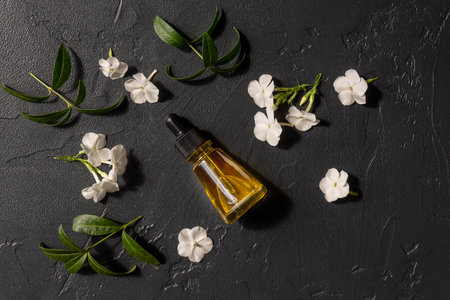 Top view of transparent bottle with beauty serum, essential oil and vitamins on dark background with white flowers and green leaves. Home spa concept.の写真素材