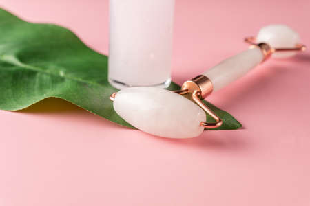 Face roller and serum dropper bottle on pink background with green leaf. Home spa proceduresの写真素材