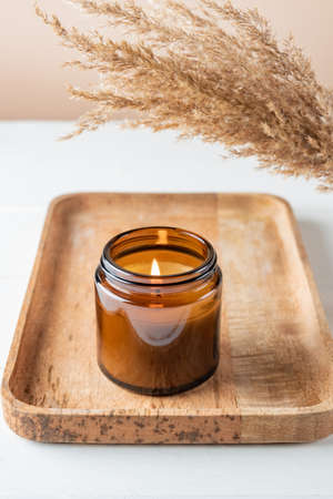 Cozy burning candle in brown glass jar, winter home interior decor on wooden tray with dry reed bouquet. Eco styleの写真素材